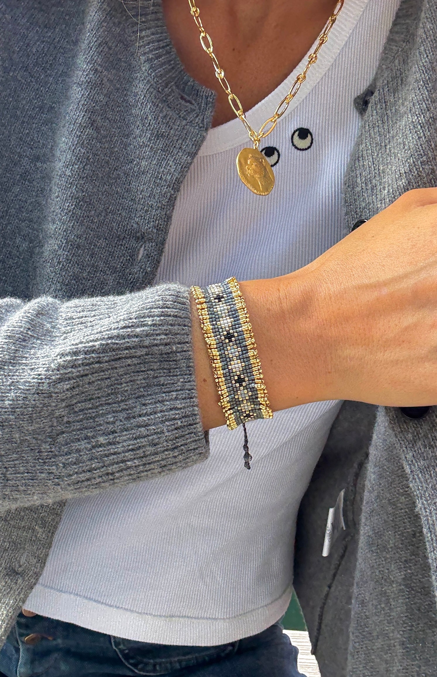 Person wearing a gold necklace and bracelet with a gray cardigan and white shirt.