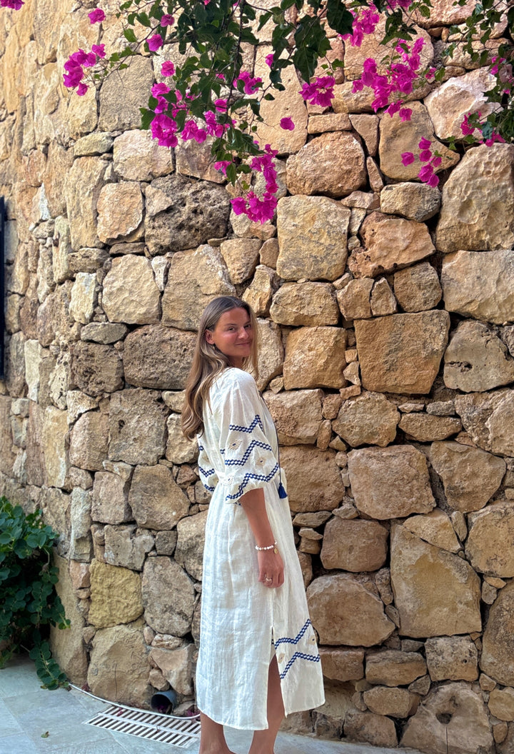 A woman wearing a white bohemian-style midi dress with blue geometric embroidery stands in front of a rustic stone wall with blooming bougainvillea flowers.