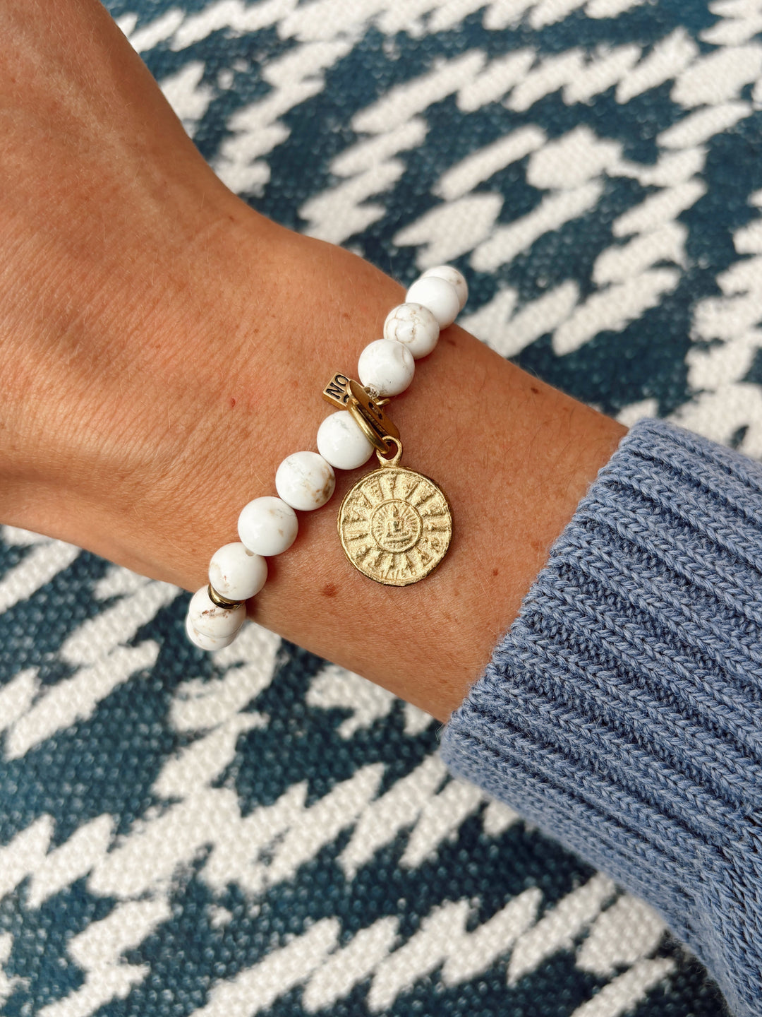 A close-up of a person's wrist wearing a beaded bracelet with white marbled beads and a gold-toned circular charm, paired with a blue ribbed sweater cuff against a navy and white patterned knit background.