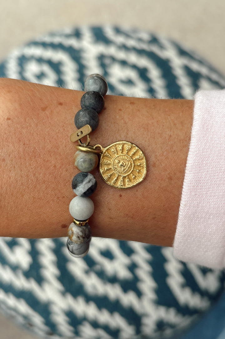 A close-up of a beaded bracelet worn on a wrist, featuring gray and white marbled beads, a small gold charm, and a round gold sun pendant. The background shows a patterned blue and white cushion and a light-colored floor.