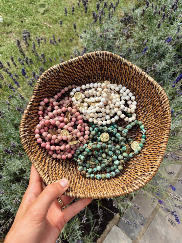 A hand holds a round woven basket outdoors, displaying three coiled beaded bracelets in pink, cream, and green with gold-toned medallion charms. Lavender plants and a stone path are blurred in the background.