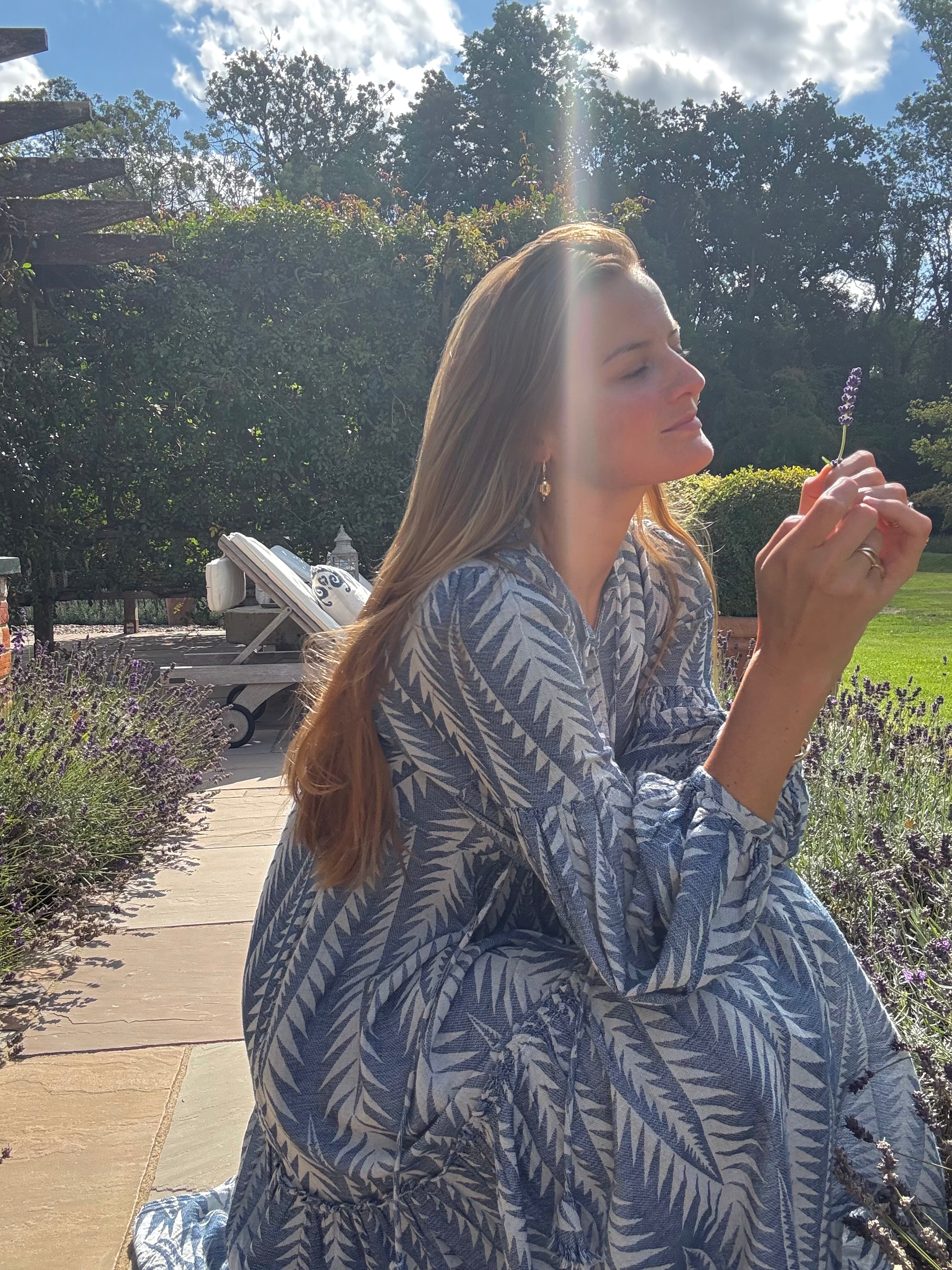 Woman in a blue and white patterned dress holding a lavender flower outdoors.