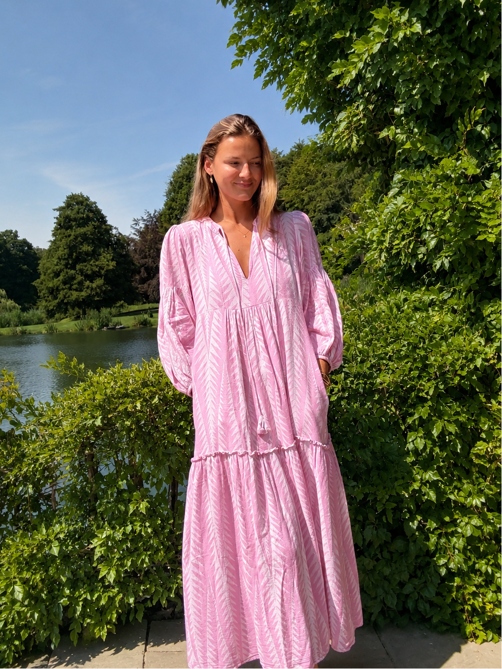 A woman wearing a light pink tiered maxi dress with a white chevron pattern stands outdoors in front of lush green trees and a lake under a blue sky.